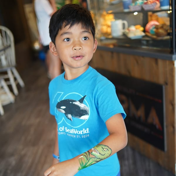 A young boy in a blue SeaWorld shirt with a dinosaur arm painting looks toward the right.
