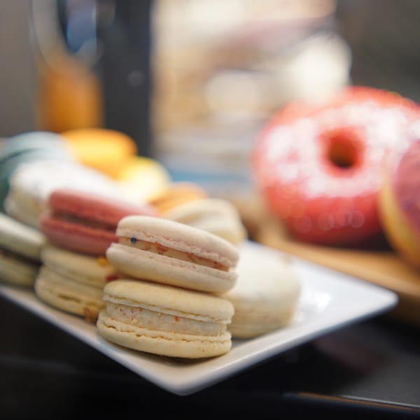 A white plate holds various colorful macarons, with a blurred pink donut in the background.