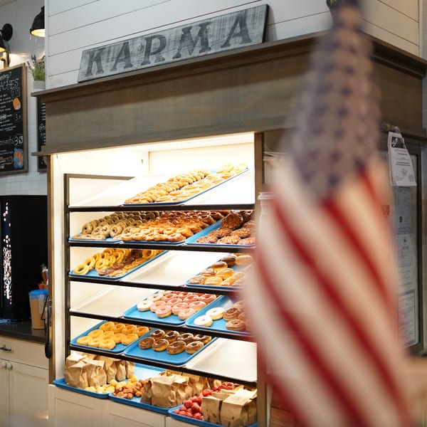 A display case at Karma Kolache & Dessert filled with various donuts and pastries, with an American flag in the foreground.