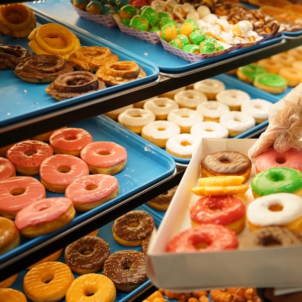 A display case filled with various colorful donuts and pastries, with a hand placing one in a box.